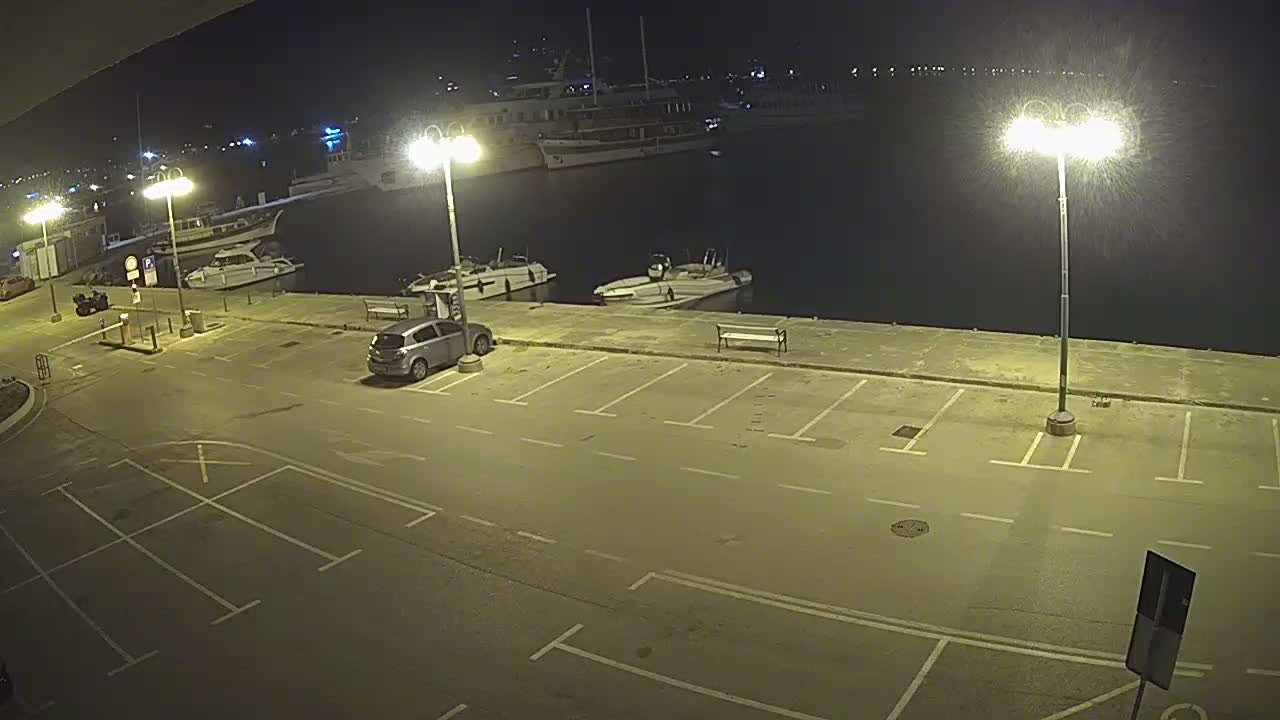 A nighttime view of an empty parking lot next to a harbor with several boats moored, illuminated by bright streetlights.