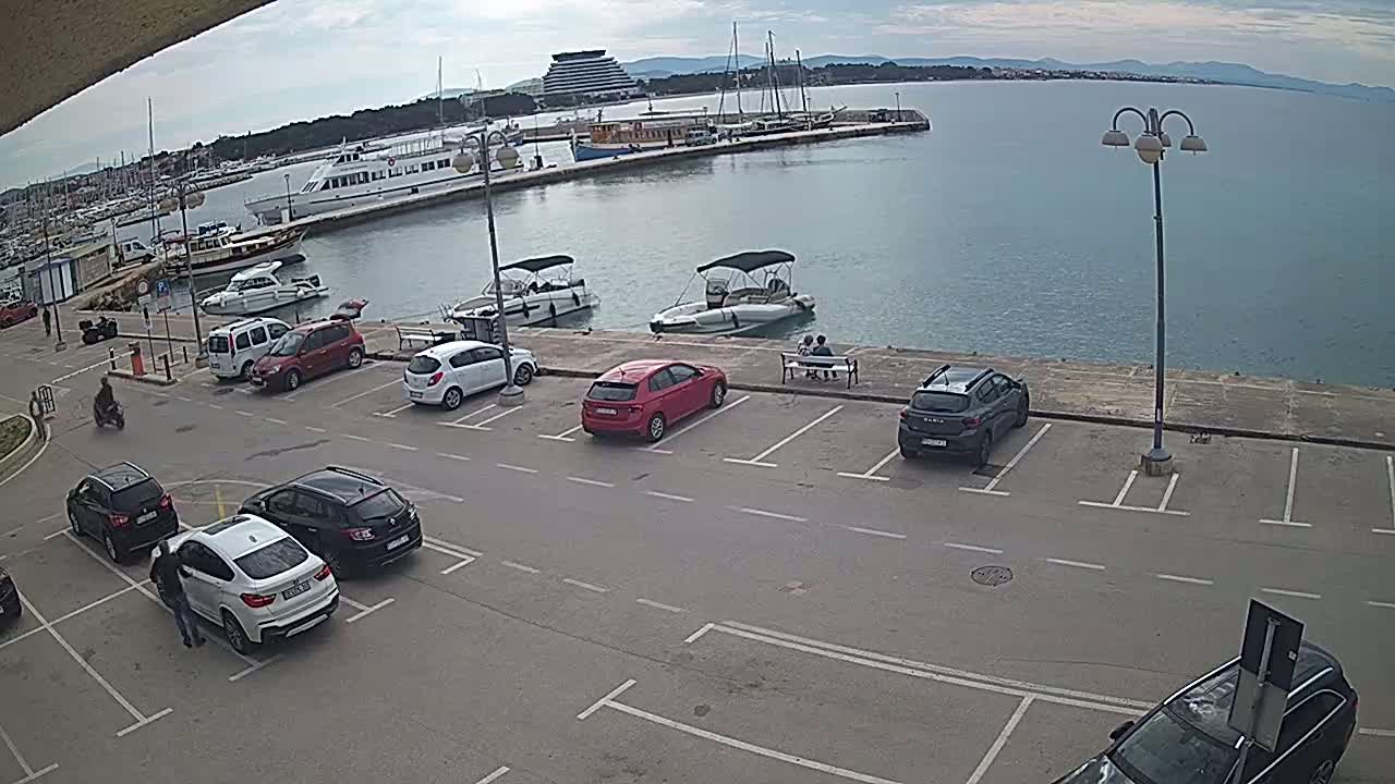 A parking lot overlooking a harbor filled with boats and a calm sea under a cloudy sky.