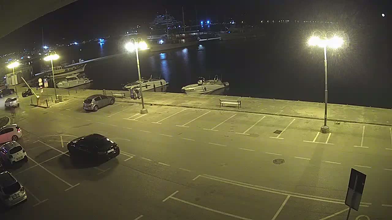 Several cars are parked in an empty lot at night next to a marina with several boats, lit by bright streetlights.