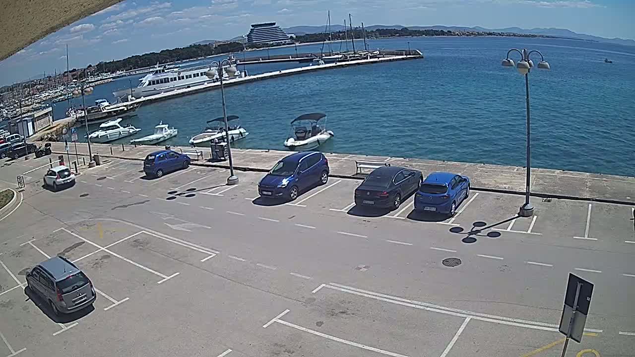 A sunny day at a harbor shows several cars parked in a lot next to a body of water with boats and a larger vessel docked.