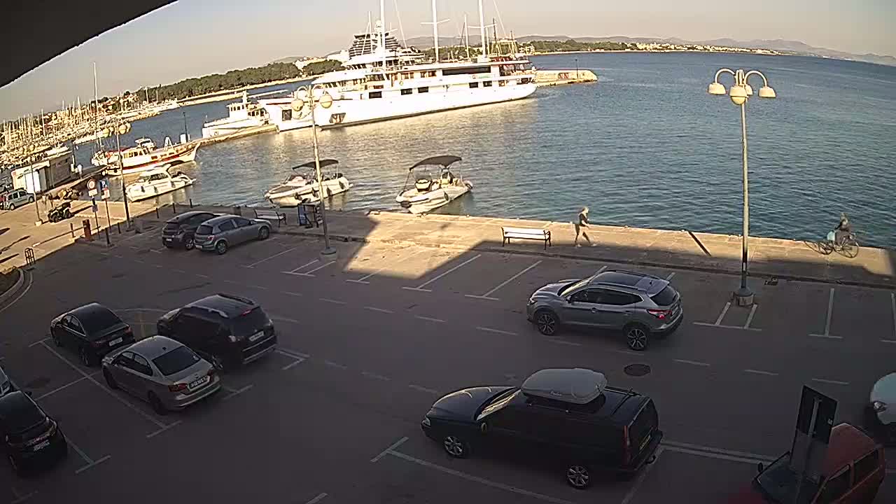 A sunny harbor scene shows several cars parked in a lot overlooking a calm body of water with many boats and a large yacht.