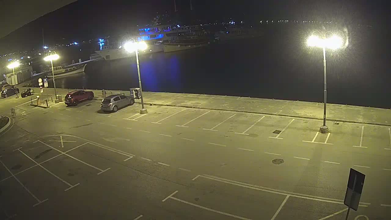 A nighttime view of an empty parking lot next to a harbor with several boats docked, illuminated by bright lights.