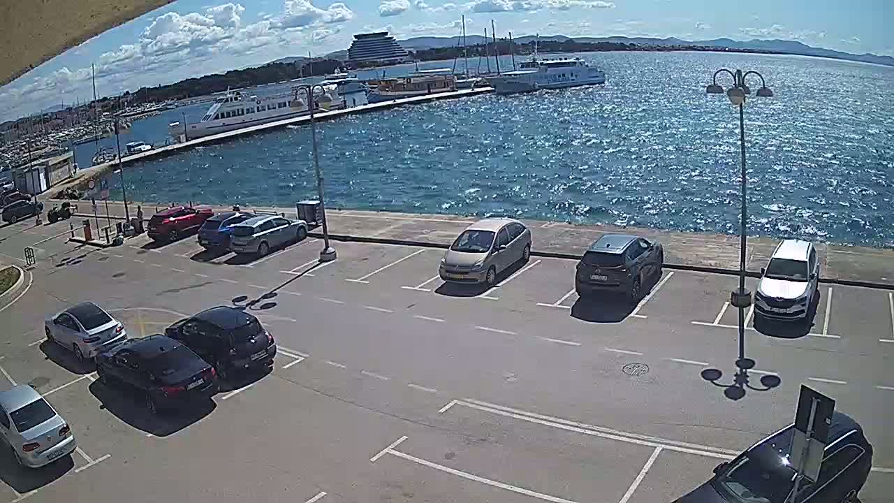 A partly sunny day shows a parking lot with several cars next to a waterfront with boats and a ferry docked.
