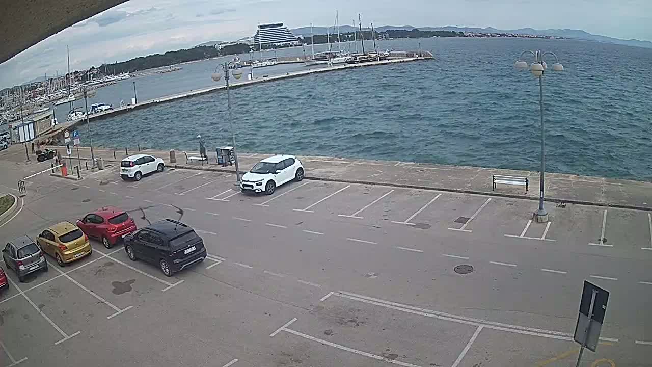 Several cars are parked in an empty lot next to a body of water with a marina and a distant building under a mostly cloudy sky.