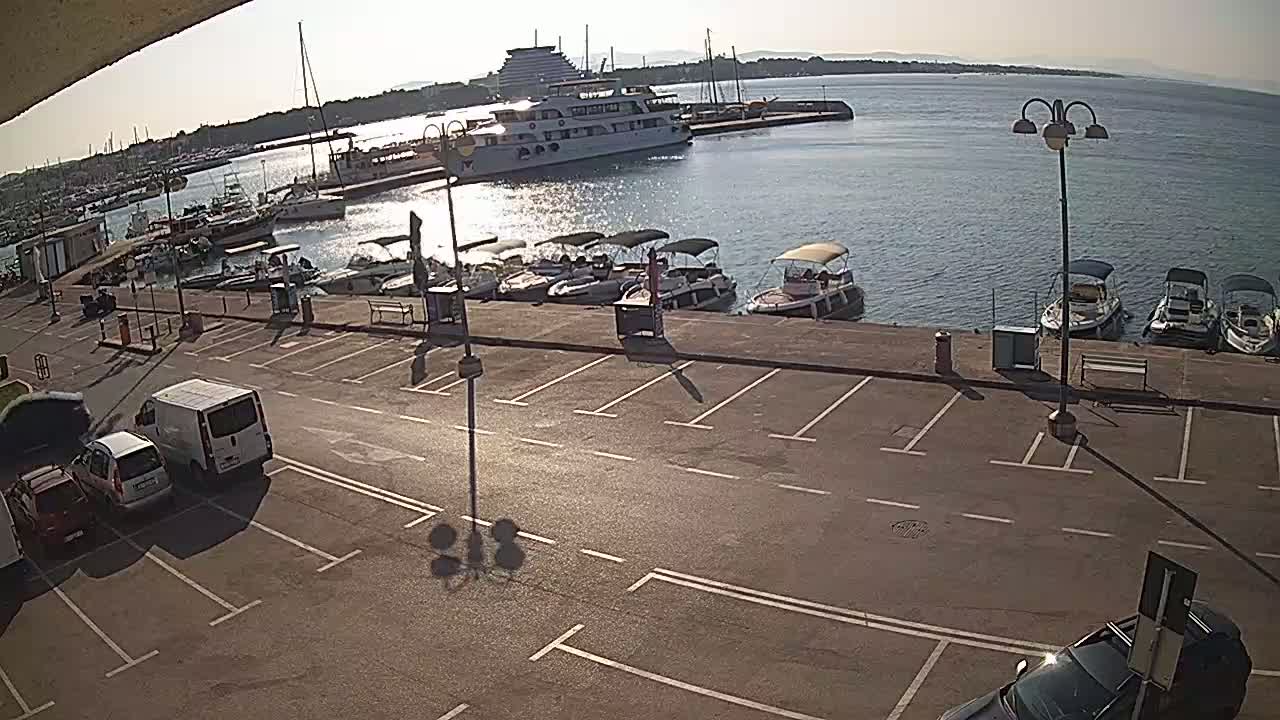 A sunny day at a marina shows a large ship and many smaller boats docked, with an empty parking lot overlooking the water.