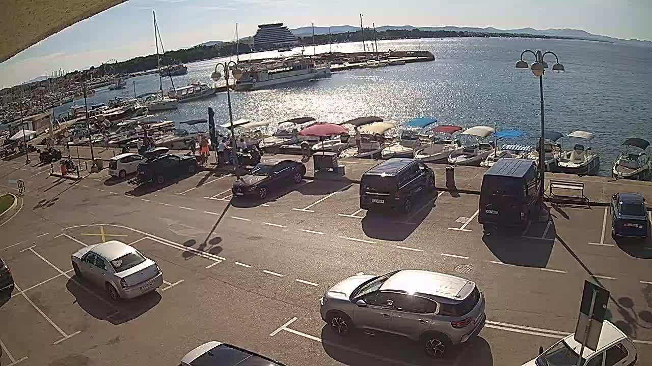 A sunny day at a harbor shows a parking lot full of cars next to a waterfront lined with docked boats and a large body of water.