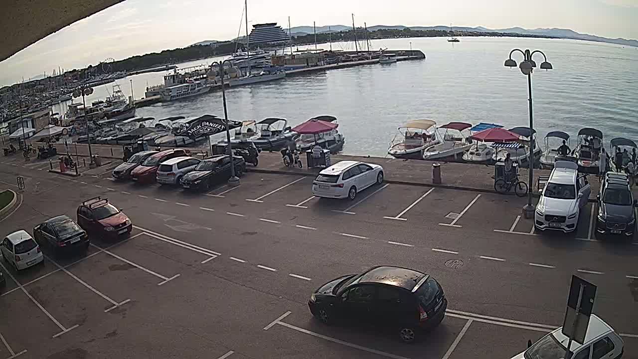 A harbor filled with boats is viewed from a parking lot on a mostly sunny day, with cars parked along the waterfront.