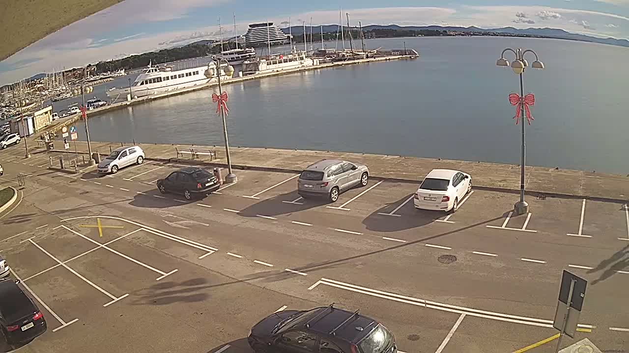 An elevated view reveals a bustling harbor on a sunny day, showcasing numerous boats docked alongside a pier with a large white vessel, several cars parked in an adjacent lot featuring lampposts adorned with red bows, and distant mountains under a blue sky with scattered clouds.
