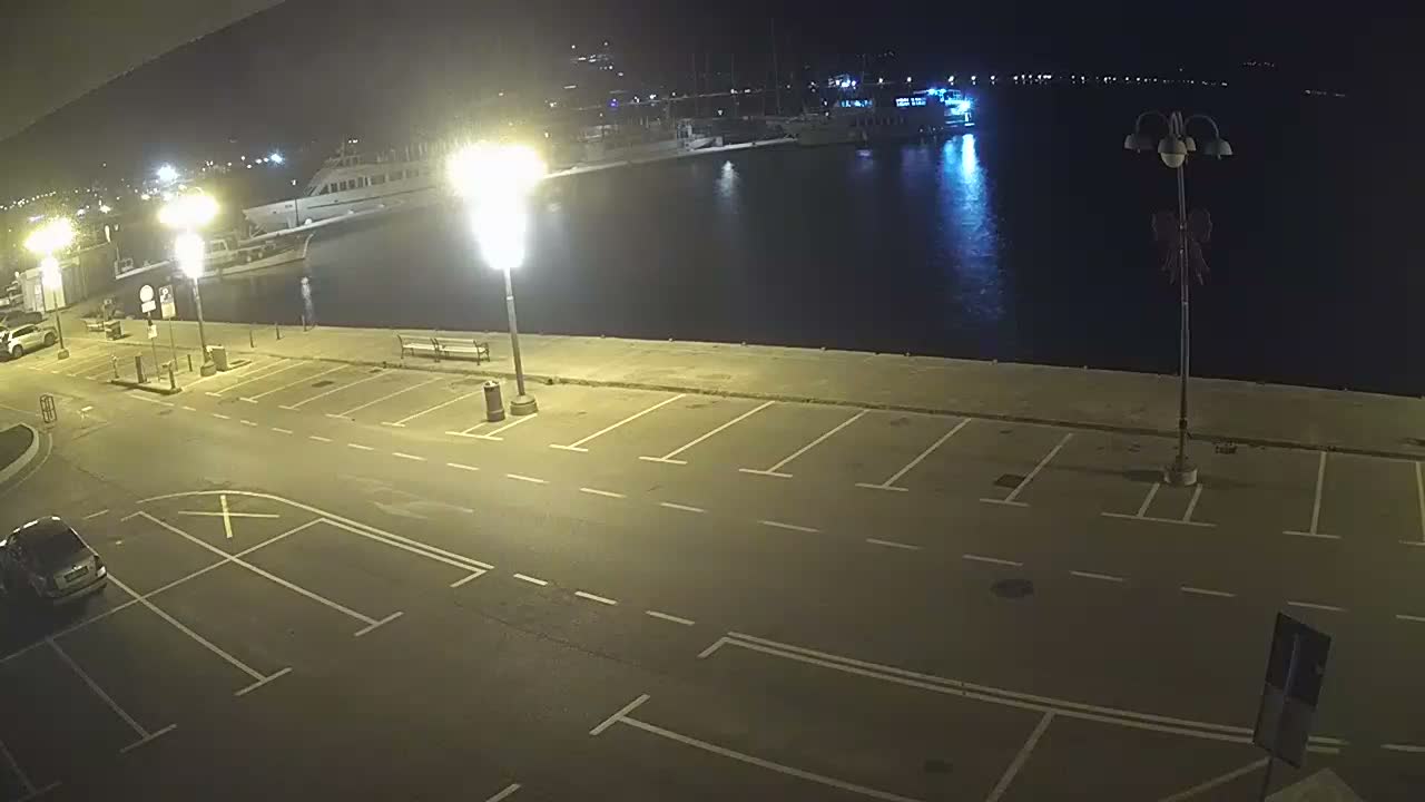 A clear night reveals a brightly illuminated waterfront with a sparse parking lot, several boats docked in the calm marina, and distant lights reflecting on the water.
