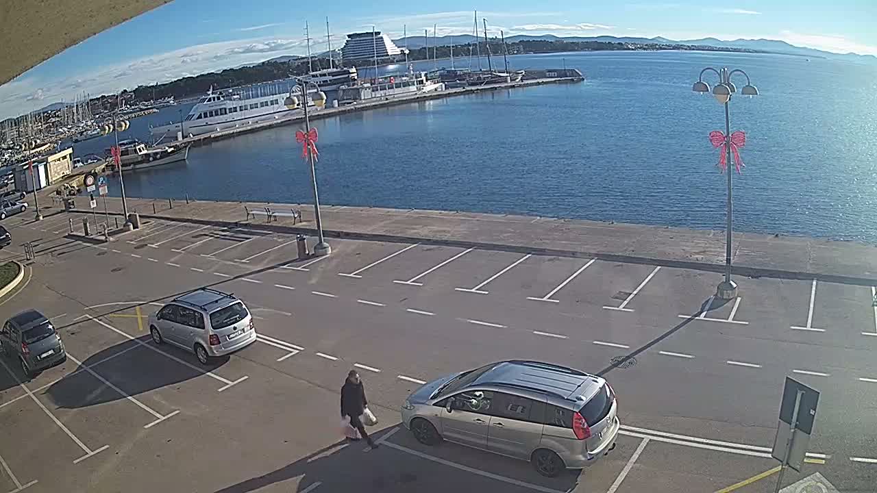 A clear, sunny day illuminates a waterfront scene featuring a busy parking lot with cars and a person walking past decorated streetlights, overlooking a harbor bustling with various boats and a large ship, leading out to a calm blue sea and distant mountains.