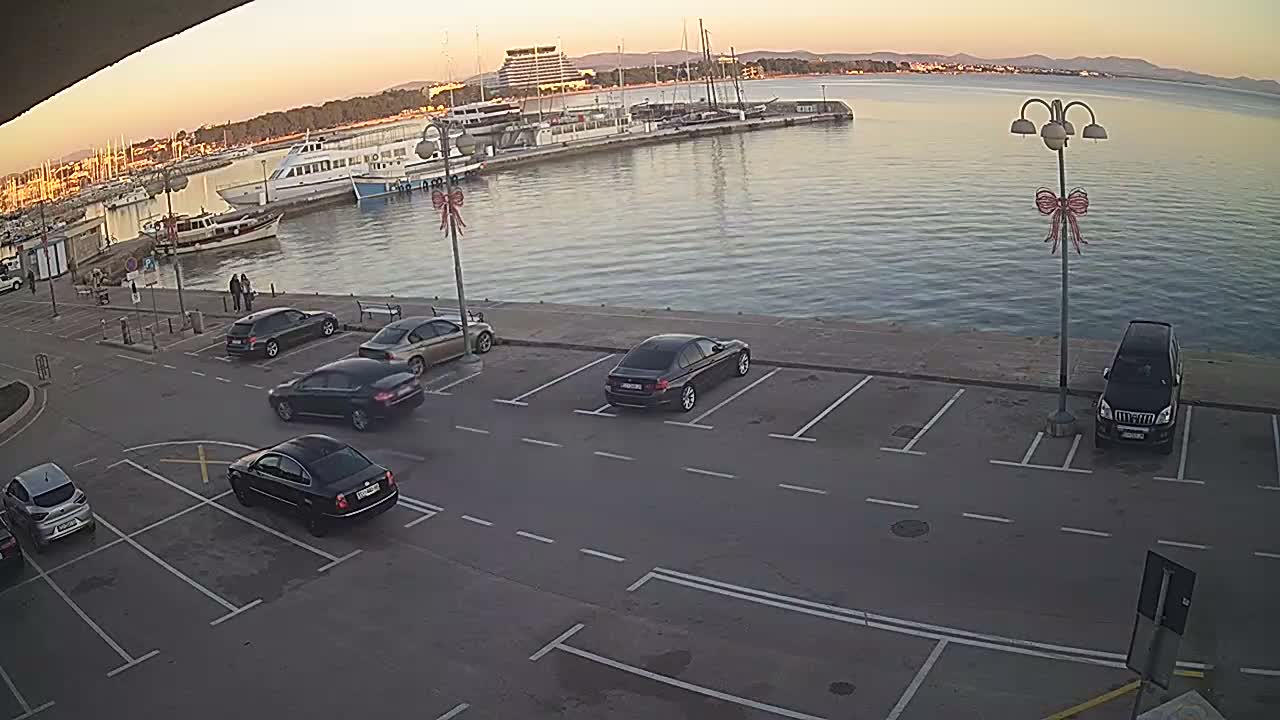 An elevated view captures a marina filled with boats, a waterfront parking lot with numerous cars, and a prominent round building on the distant shore, all beneath a partly cloudy sky.