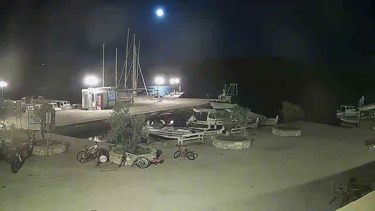 A moonlit night scene shows a harbor with several boats docked, along with bicycles and small vehicles parked on the adjacent land.