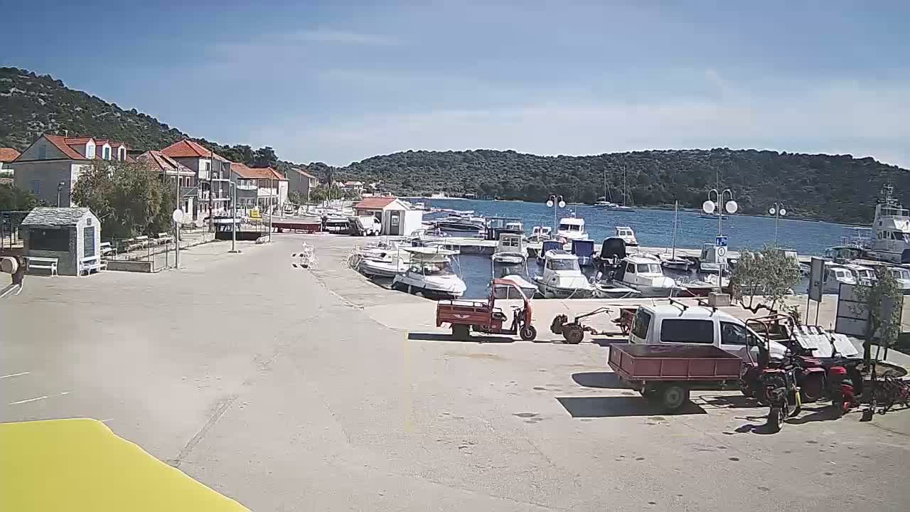 A sunny day at a harbor shows several boats docked, along with small vehicles and buildings nearby.