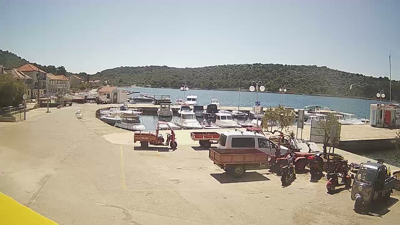 Under a clear sky, a harbor is filled with numerous small boats and is surrounded by buildings and greenery; several small vehicles and motorized scooters are parked in the foreground.