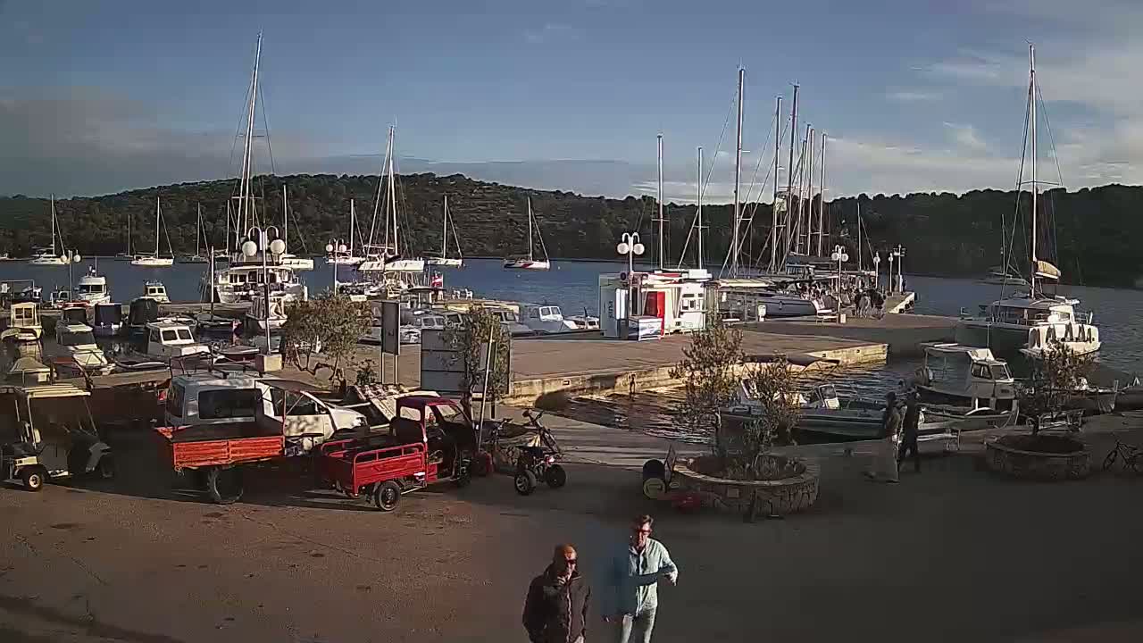A marina filled with sailboats and motorboats is seen on a sunny day, with several small vehicles and two people walking in the foreground.
