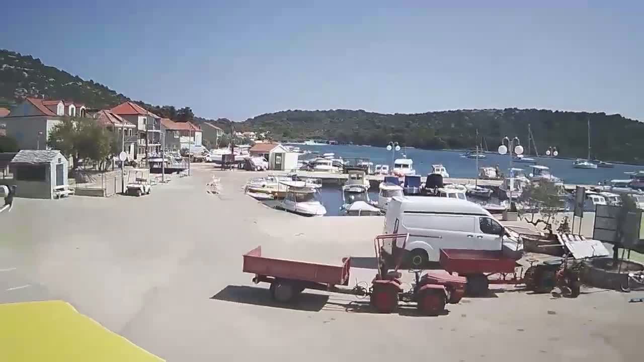 A sunny day reveals a small harbor filled with boats, adjacent to a village with buildings and parked vehicles, including a van and small tractor with a trailer.