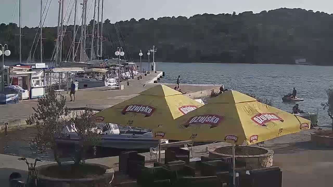 A marina with numerous sailboats and a few people, some yellow umbrellas, and a jet ski in calm water under a sunny sky.