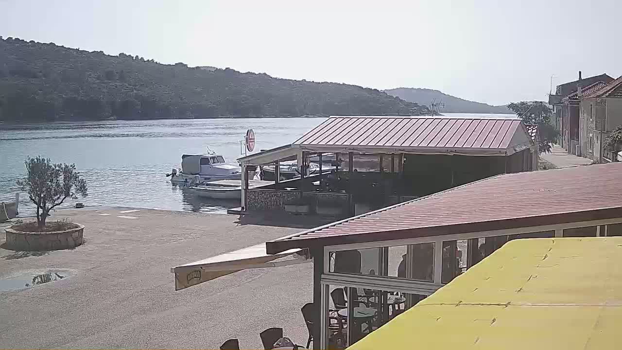 A calm, sunny day at a waterfront location shows boats docked near an outdoor restaurant with a red roof, overlooking a bay surrounded by green hills.