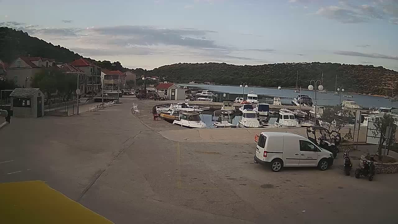 A small harbor with numerous boats docked, a few buildings, and a parking lot with a white van and scooters, under a partly cloudy sky.