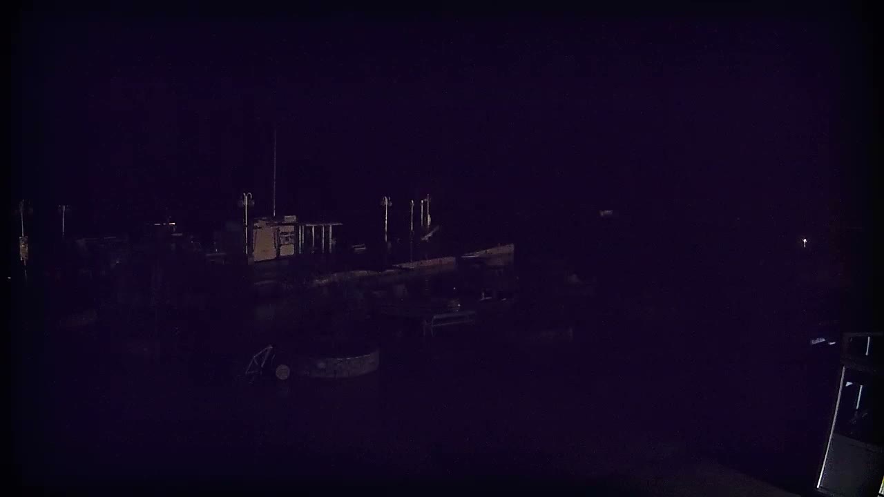 On a partly cloudy day, a harbor scene features several boats docked at piers, forested hills across a calm bay, and a dark utility vehicle and a red three-wheeled vehicle on the paved waterfront.