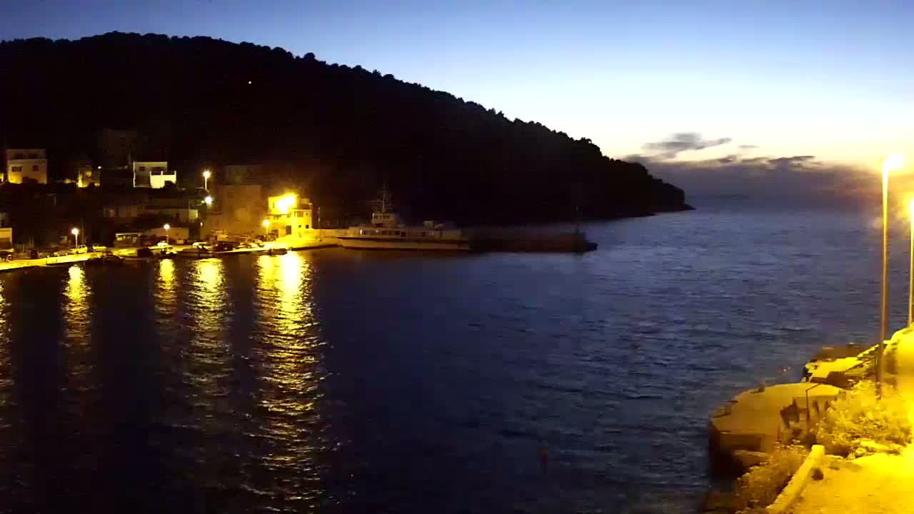 A nighttime view of a calm harbor with a small town nestled against a dark hill, illuminated by streetlights reflecting on the water.