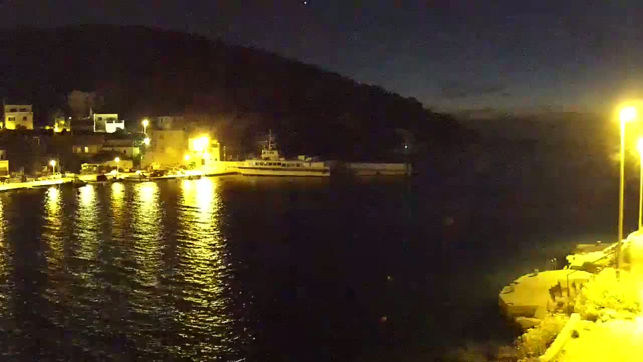 A nighttime view of a harbor with a docked ship, illuminated buildings on a hillside, and calm water reflecting the lights.