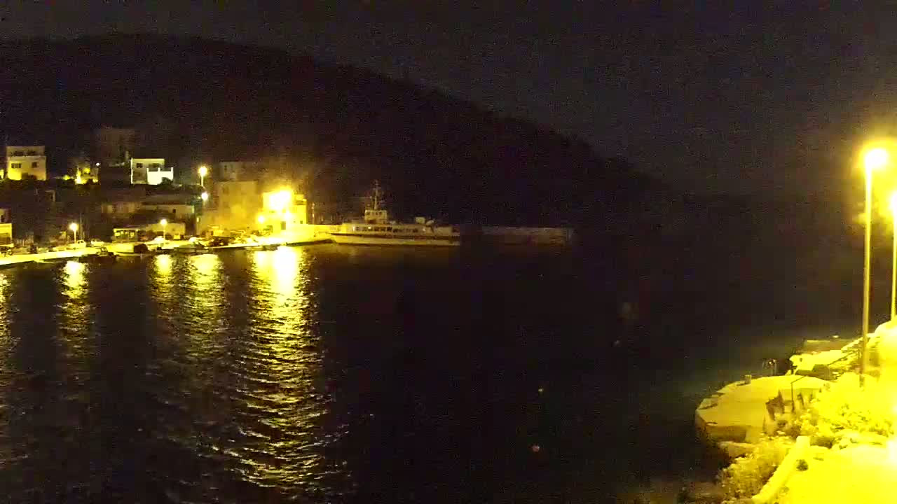 A nighttime view of a harbor with a boat docked, illuminated by streetlights, beside a small village nestled against a dark hillside.
