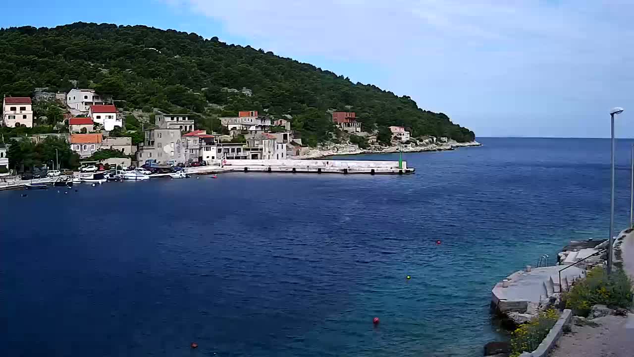 A small village with red-roofed houses clusters along a calm, blue sea on a partly cloudy day, nestled against a verdant hillside.