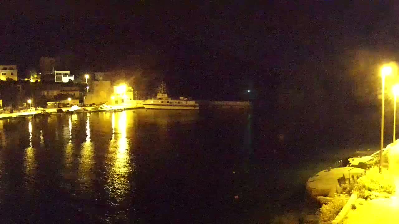 A nighttime view of a harbor with a ship docked near illuminated buildings, reflecting lights on calm water.