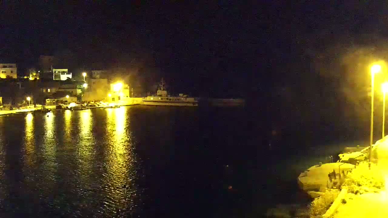 A nighttime harbor scene shows a calm body of water reflecting the lights from a small town on a hill, with a ship moored in the harbor.