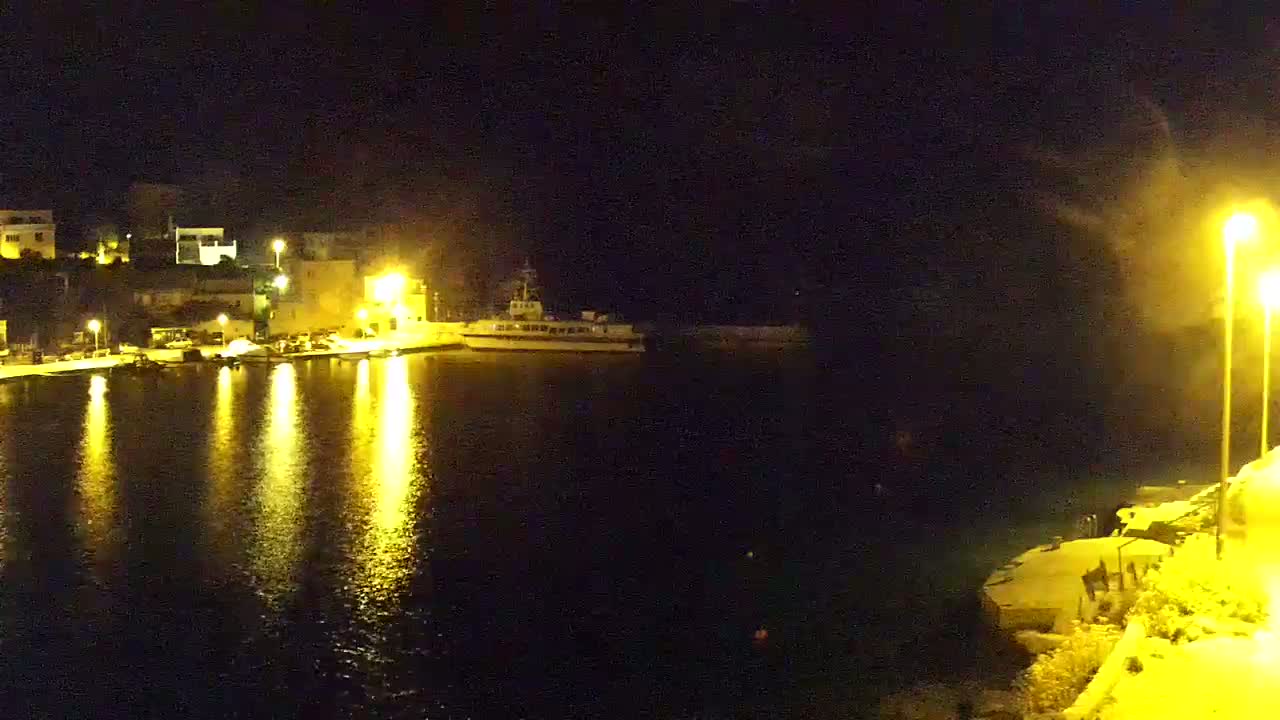 A nighttime view of a harbor with a docked ship, illuminated buildings along the shore, and calm water reflecting the lights.