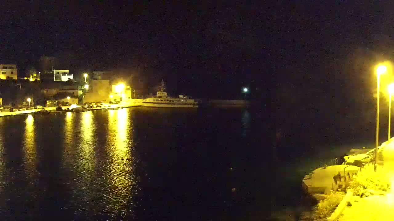 A nighttime harbor scene shows a docked ship and illuminated buildings reflected in calm water under bright lights.