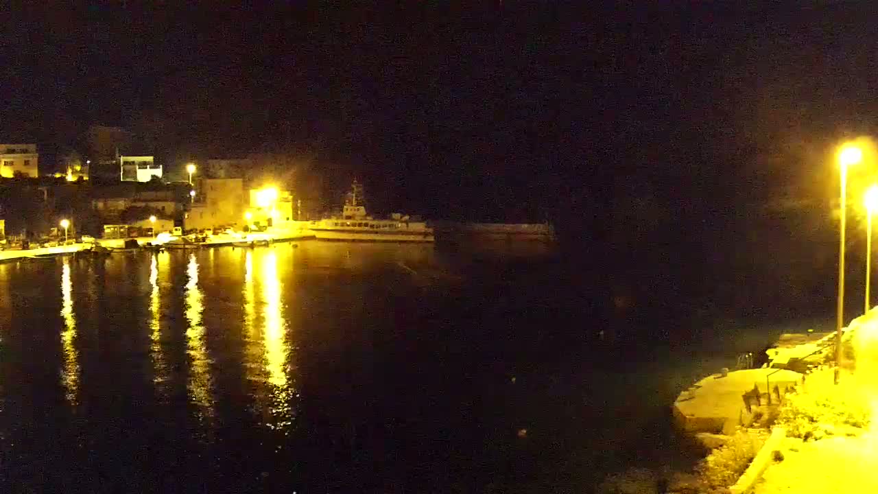 A nighttime view of a harbor with buildings illuminated on one side, a ship docked in the water, and the reflections of lights shimmering on the dark water.