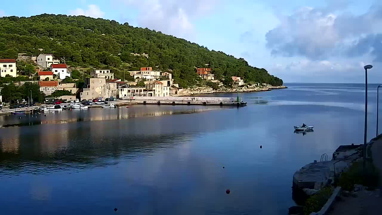 A calm, partly cloudy day shows a small village nestled on a hillside overlooking a tranquil bay with several boats moored at a harbor.