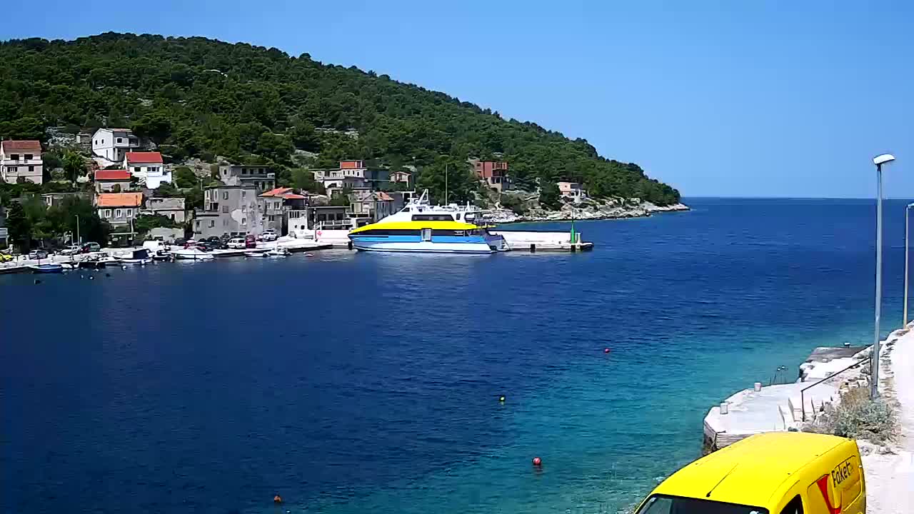 A yellow and blue ferry is docked at a harbor next to a small village nestled against a lush green hillside on a sunny day.