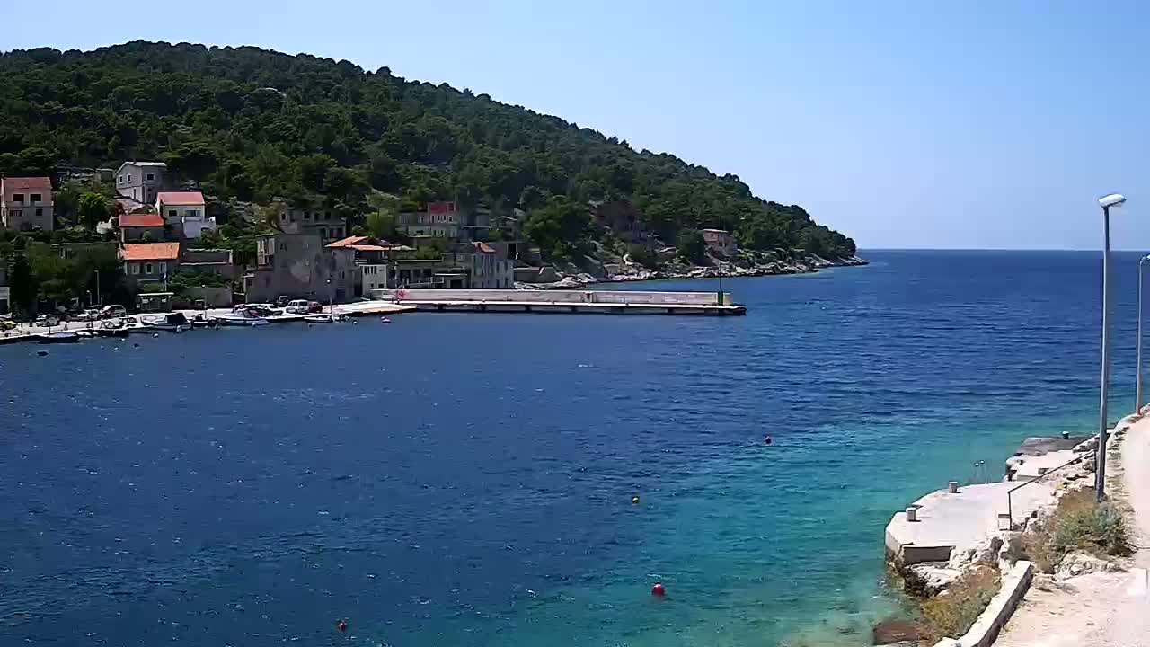 A small waterfront village nestled against a verdant hillside, with calm blue water and a clear sunny sky.