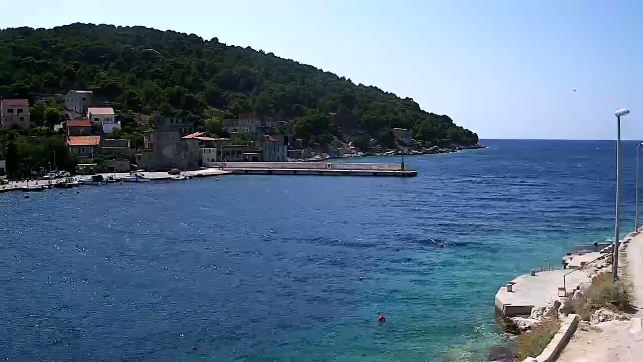 A small coastal town nestled against a lush green hill overlooks a calm, blue sea on a sunny day.