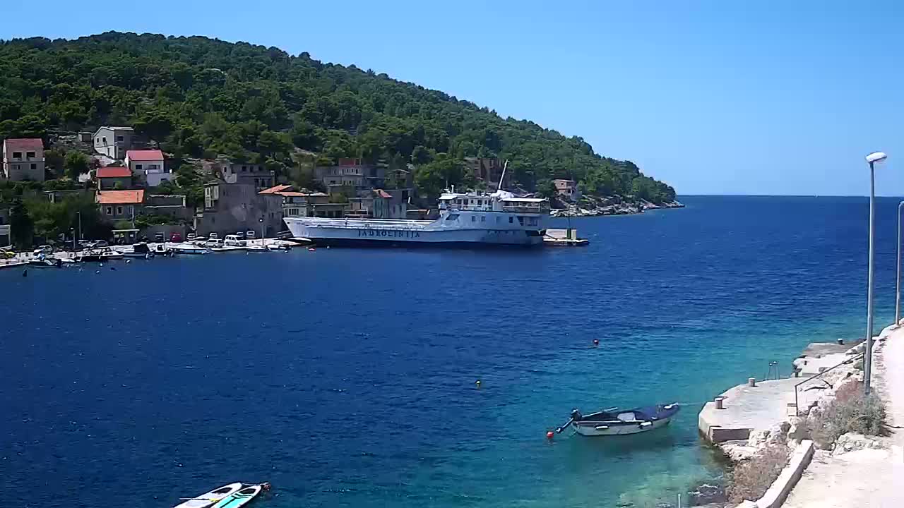 A small coastal town nestled against a lush green hill overlooks a calm, blue sea on a sunny day.