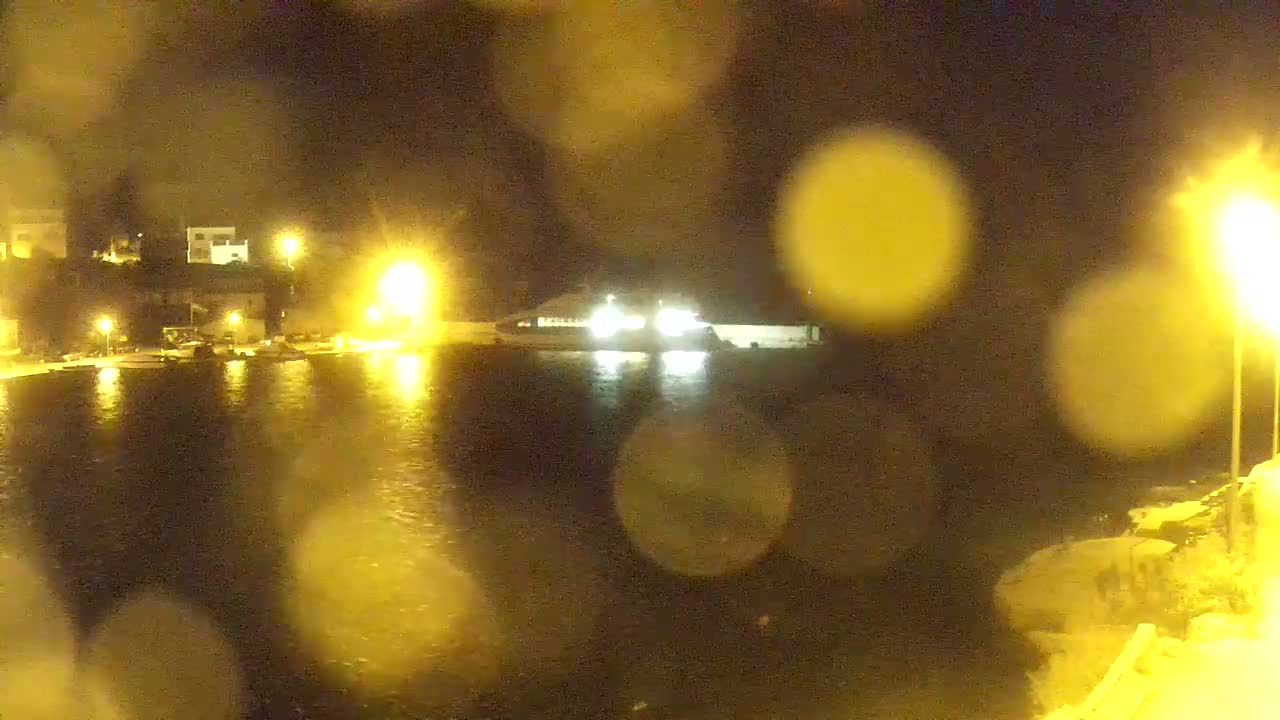 A small coastal town nestled against a lush green hill overlooks a calm, blue sea on a sunny day.