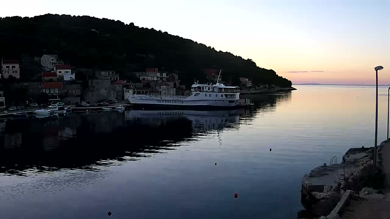 A small coastal town nestled against a lush green hill overlooks a calm, blue sea on a sunny day.
