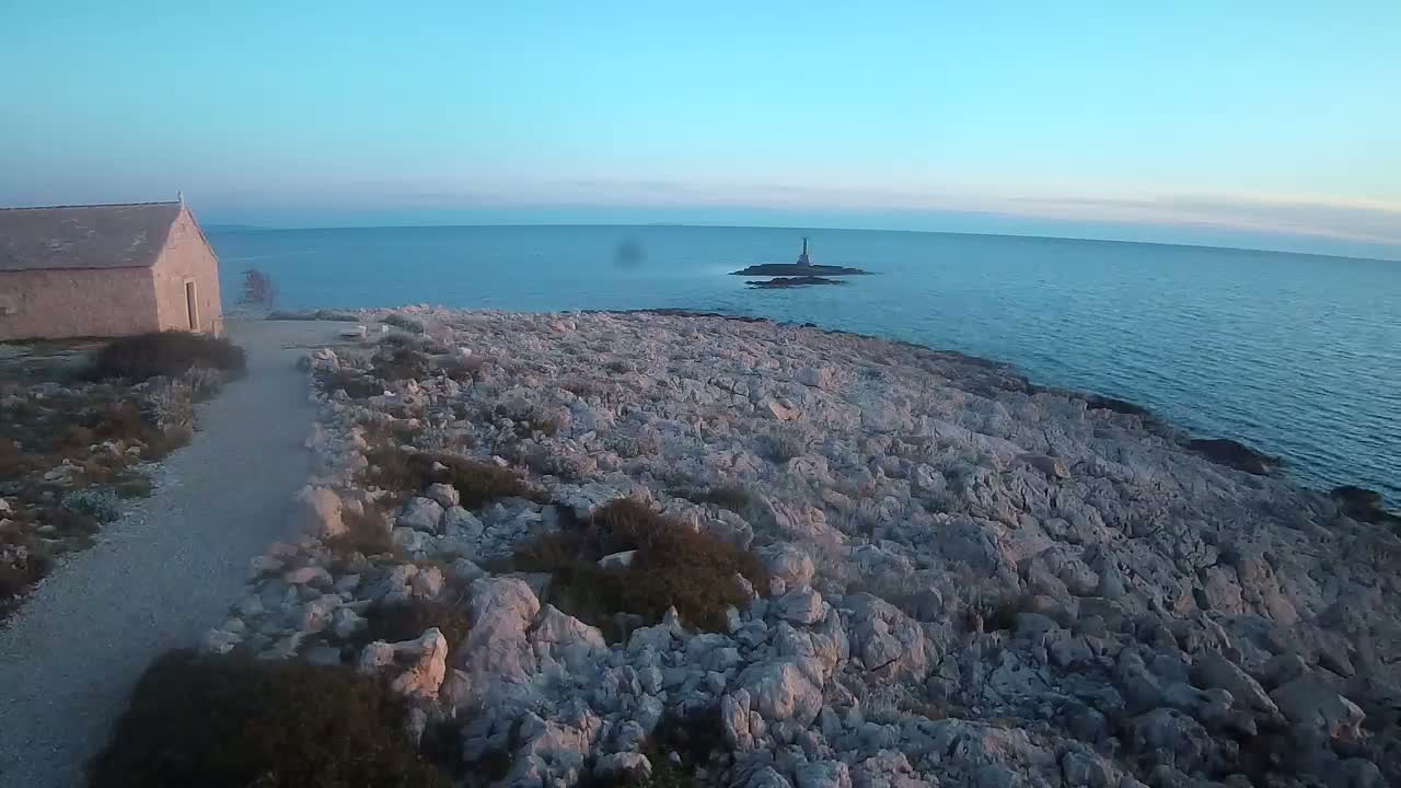 A stone building sits on a rocky shoreline overlooking a calm, blue sea and a small island with a lighthouse under a clear, twilight sky.