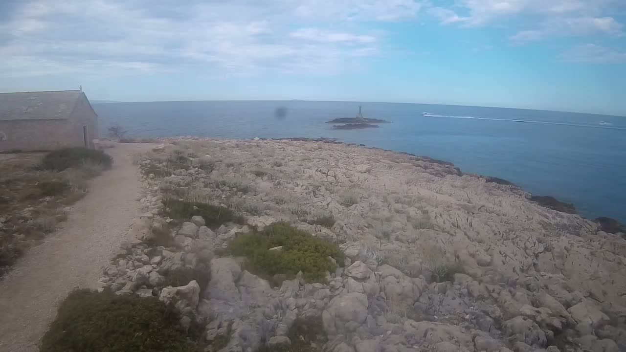 A stone building sits near a rocky, sparsely vegetated coastline overlooking a calm sea with a small island and a few boats in the distance under a partly cloudy sky.