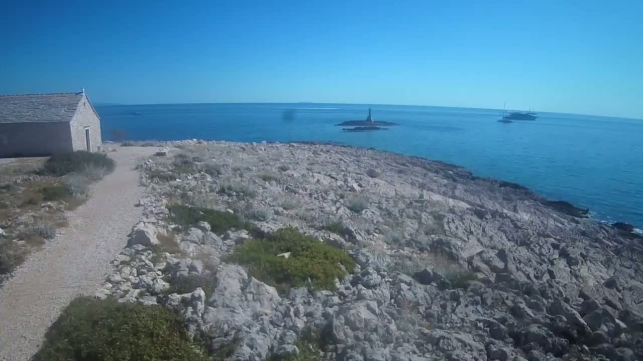 A stone building sits near a rocky, sparsely vegetated coastline overlooking a calm sea with a small island and a few boats in the distance under a partly cloudy sky.