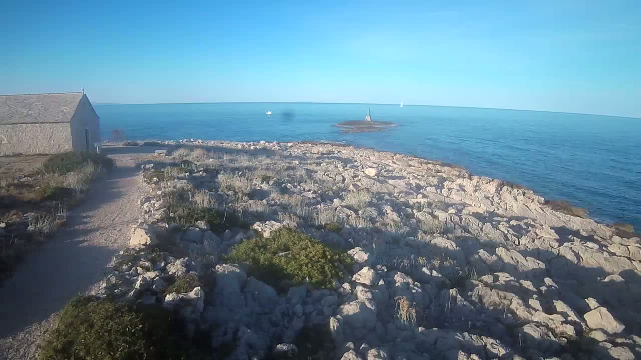 A stone building sits near a rocky, sparsely vegetated shoreline on a clear, sunny day overlooking a calm blue sea with a small island and a couple of distant sailboats visible.