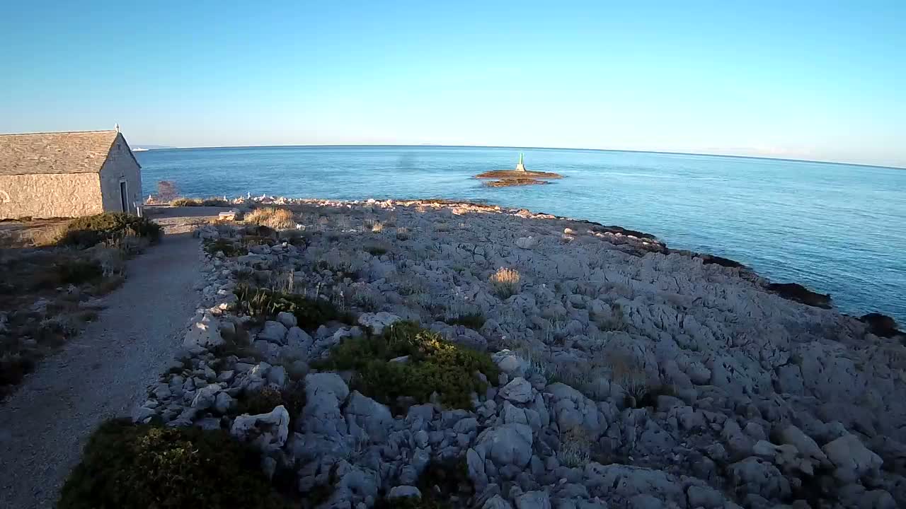 A small stone building sits near a rocky shoreline overlooking a calm blue sea with a few sailboats in the distance under a bright sunny sky.