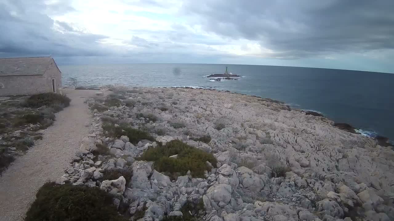 A couple walks along a rocky path near a small stone building overlooking a calm blue sea with a distant lighthouse under a clear blue sky.