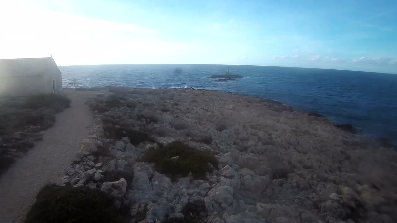 A bright, sunny day illuminates a rocky coastal landscape with a small light-colored building and a dirt path on the left, leading towards a choppy blue ocean featuring a distant lighthouse on a rocky islet.