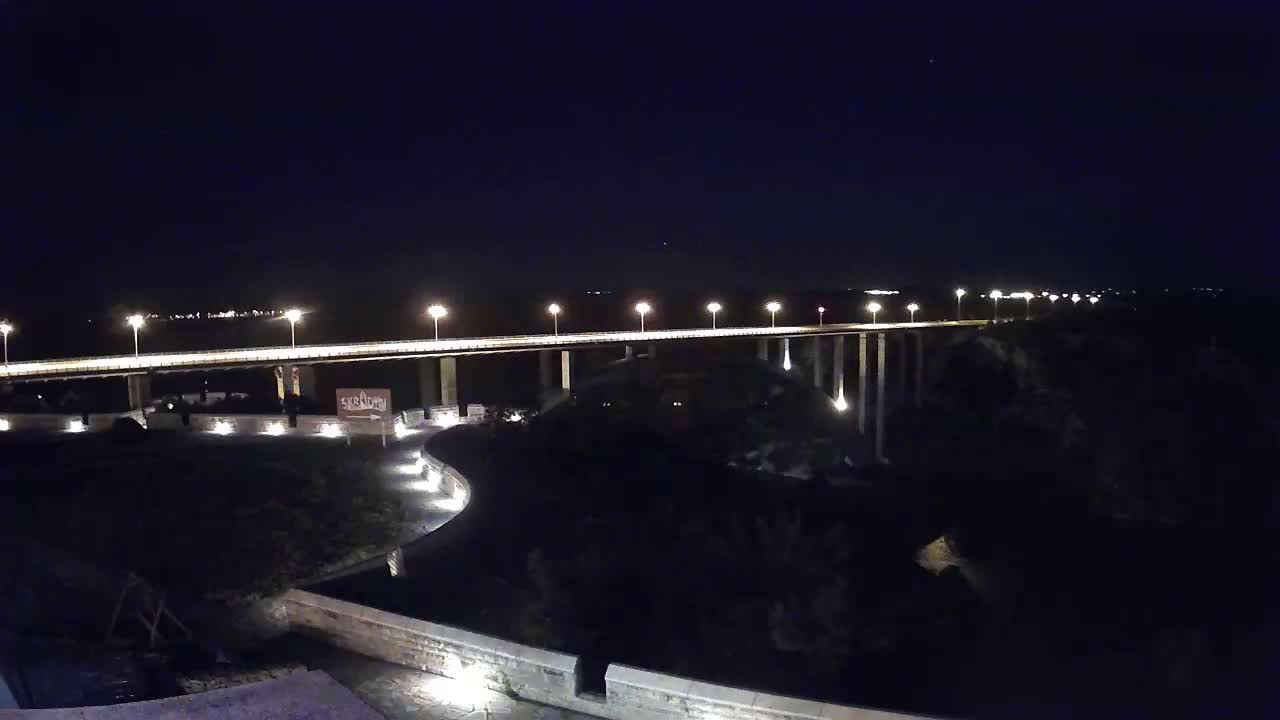 A long, brightly lit bridge spans a dark body of water at night under a clear, starlit sky.