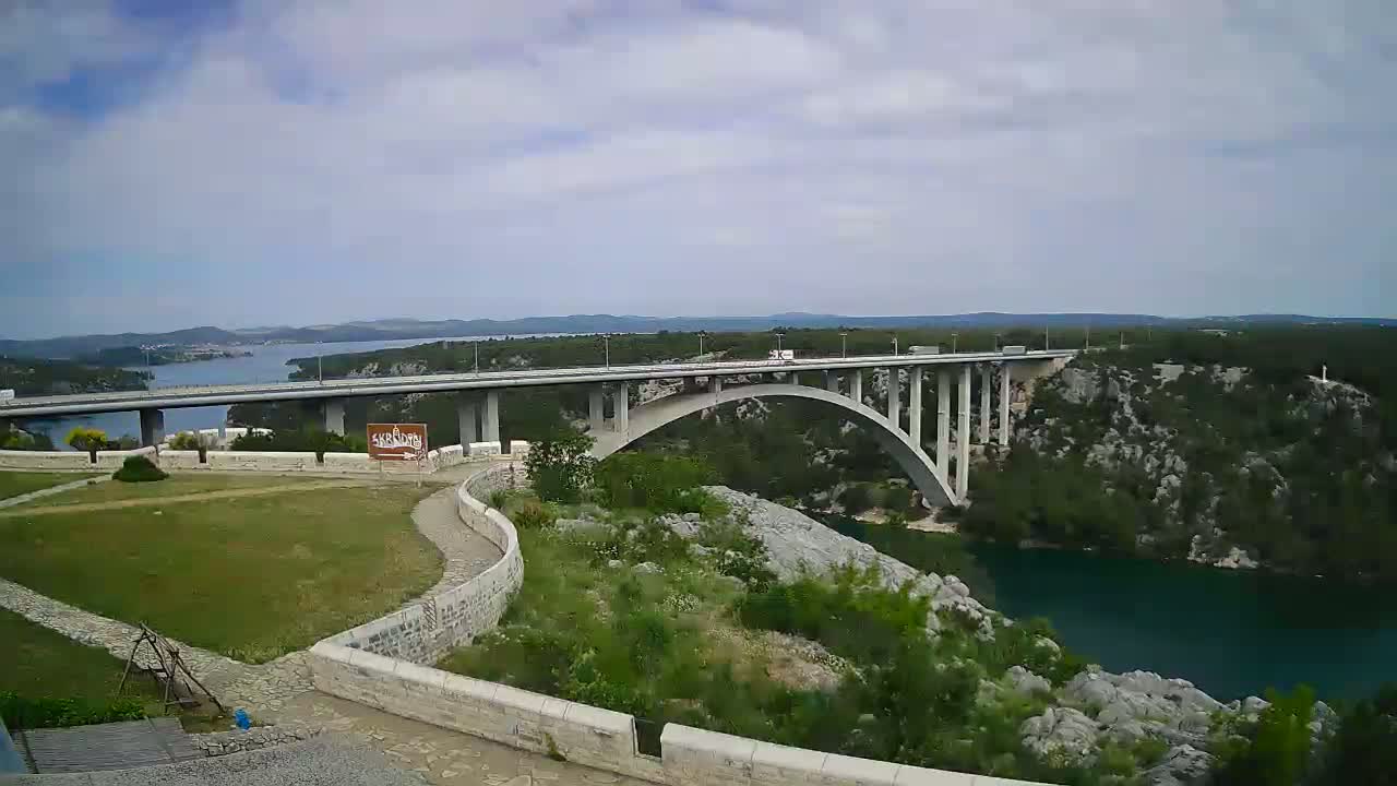 A long bridge stretches across a body of water under a partly cloudy sky, with green vegetation and rocky terrain surrounding it.