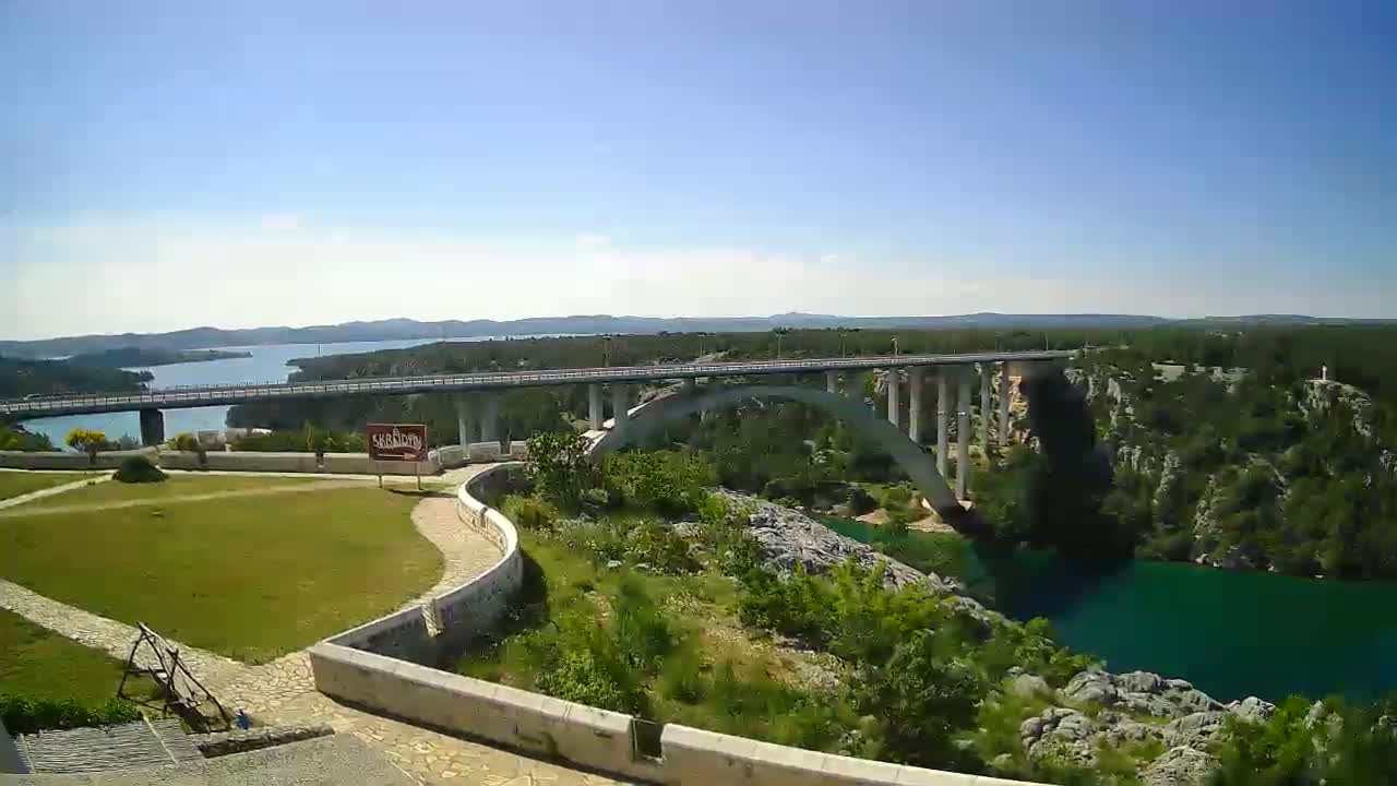 A long bridge spans a vibrant blue-green river, with lush green vegetation and a clear sunny sky visible in the background.